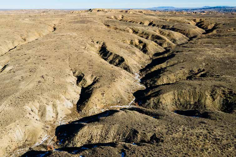 Aerial view of deep eroded ravines cutting through arid badlands terrain under clear blue sky.