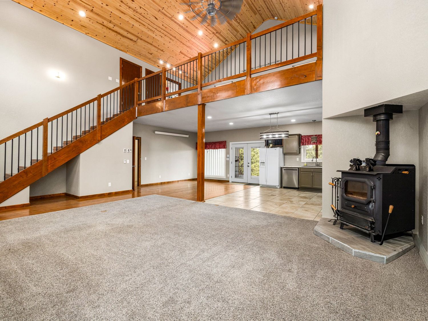 Spacious open-concept living room with vaulted wood ceiling, loft balcony, staircase, carpet flooring, and a black wood-burning stove.