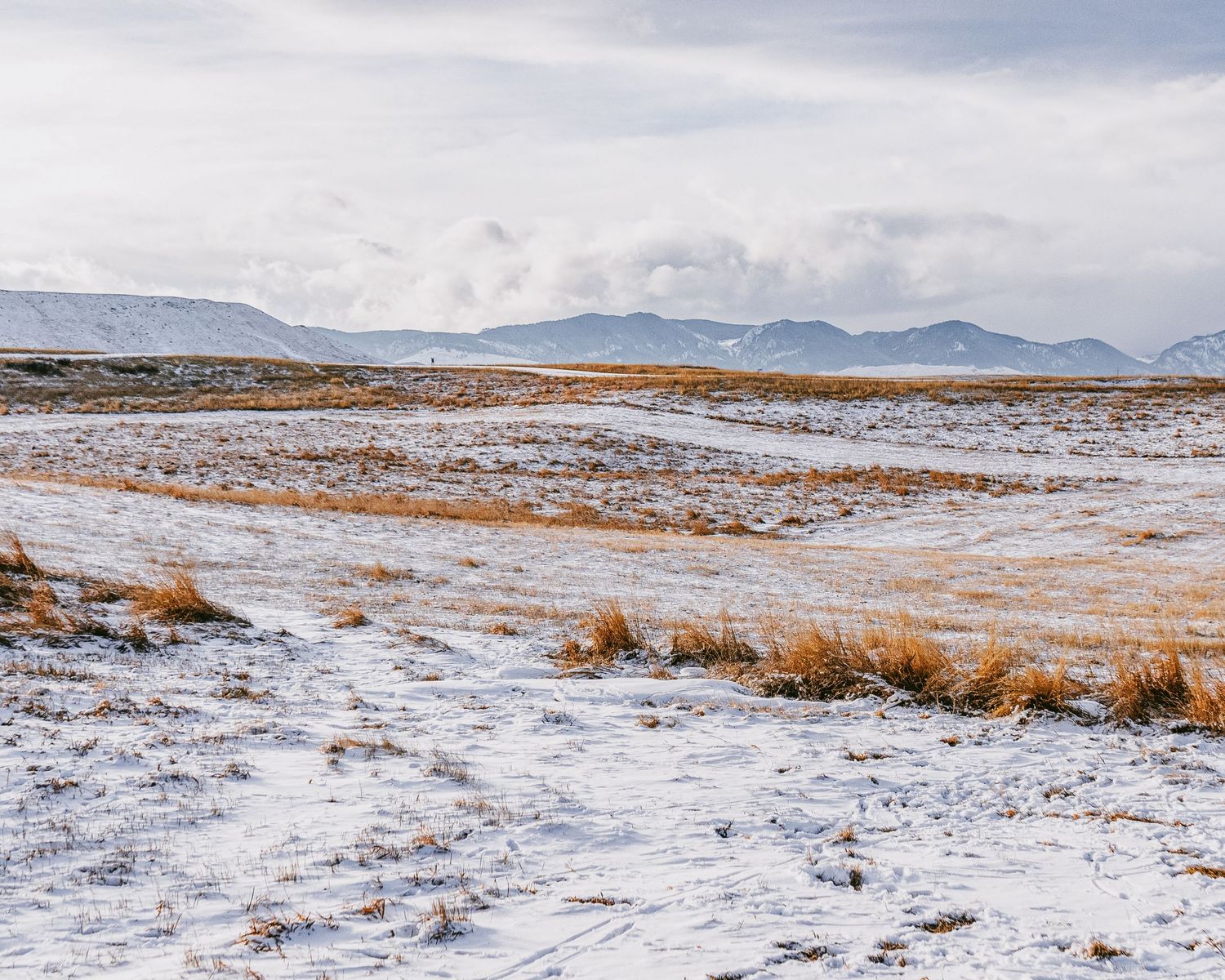 Snow-covered winter landscape with rolling hills and mountains in the distance under cloudy skies.