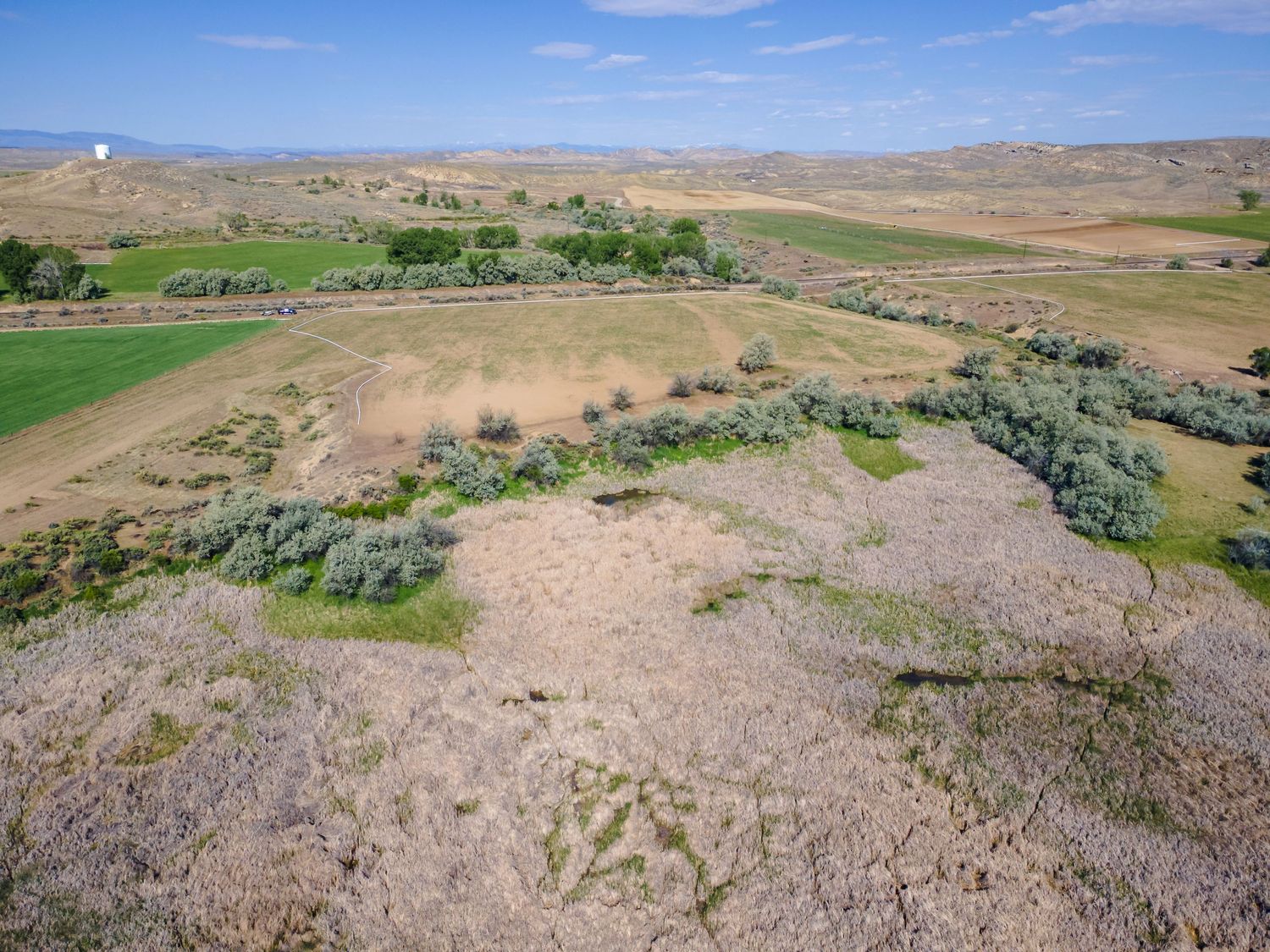 Aerial view of vast rural farmland with rolling hills, sagebrush, green fields, and a water tower under a partly cloudy sky.