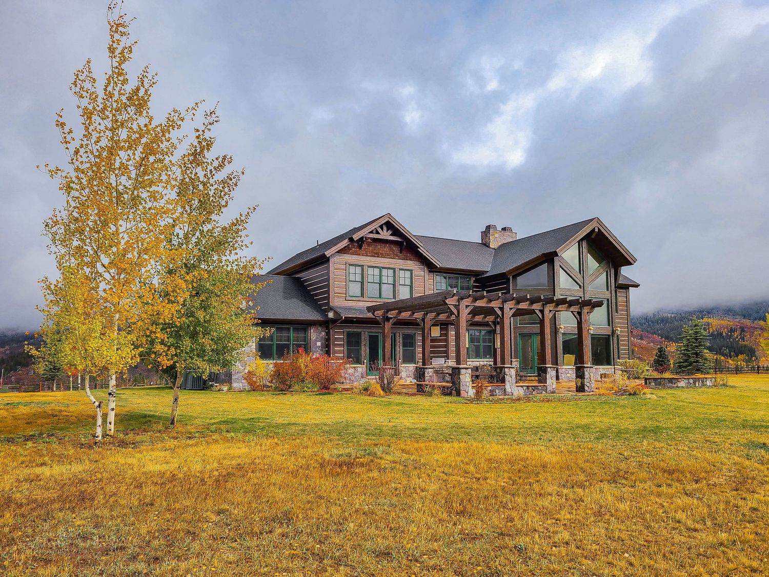 Rustic mountain home with log accents, covered porch, surrounded by golden autumn foliage and open green meadows under cloudy skies.