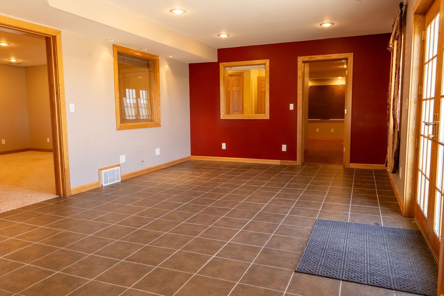 Spacious empty room with dark tile flooring, red accent wall, wood-trimmed windows, and recessed lighting.
