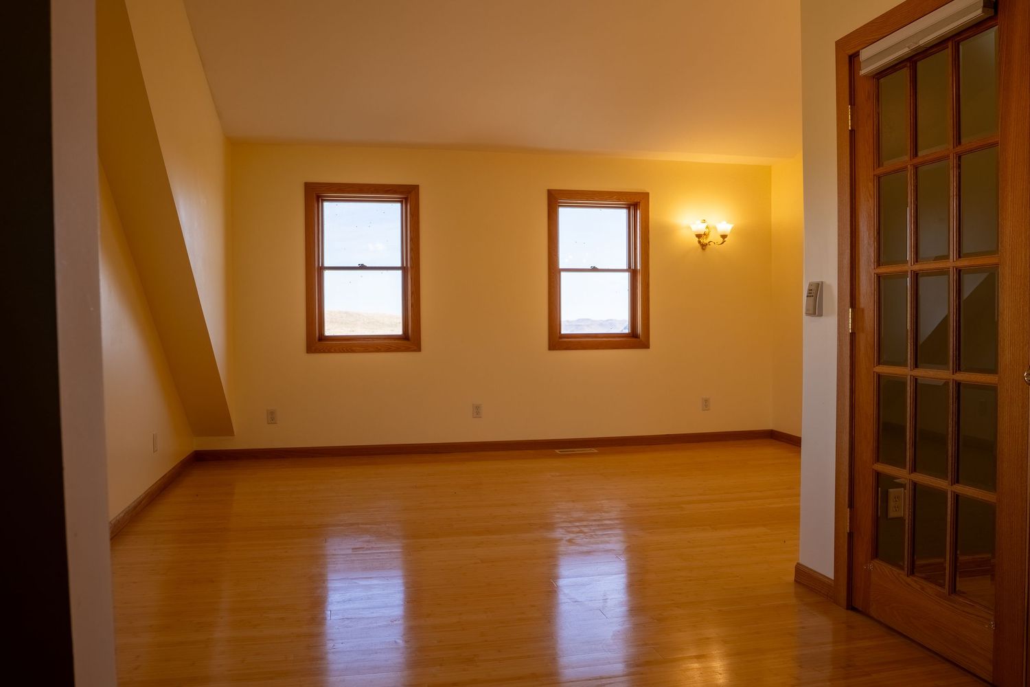 Empty room with hardwood floors, two windows, yellow walls, and a French door in a cozy attic space.