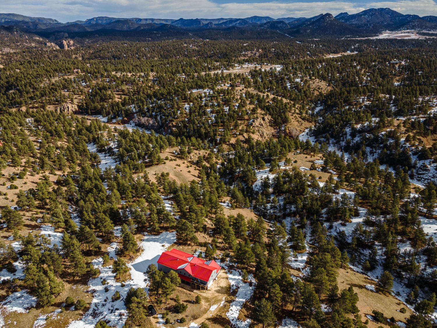 Drone shot of a red-roofed house nestled among pine trees and snow patches in mountainous terrain.
