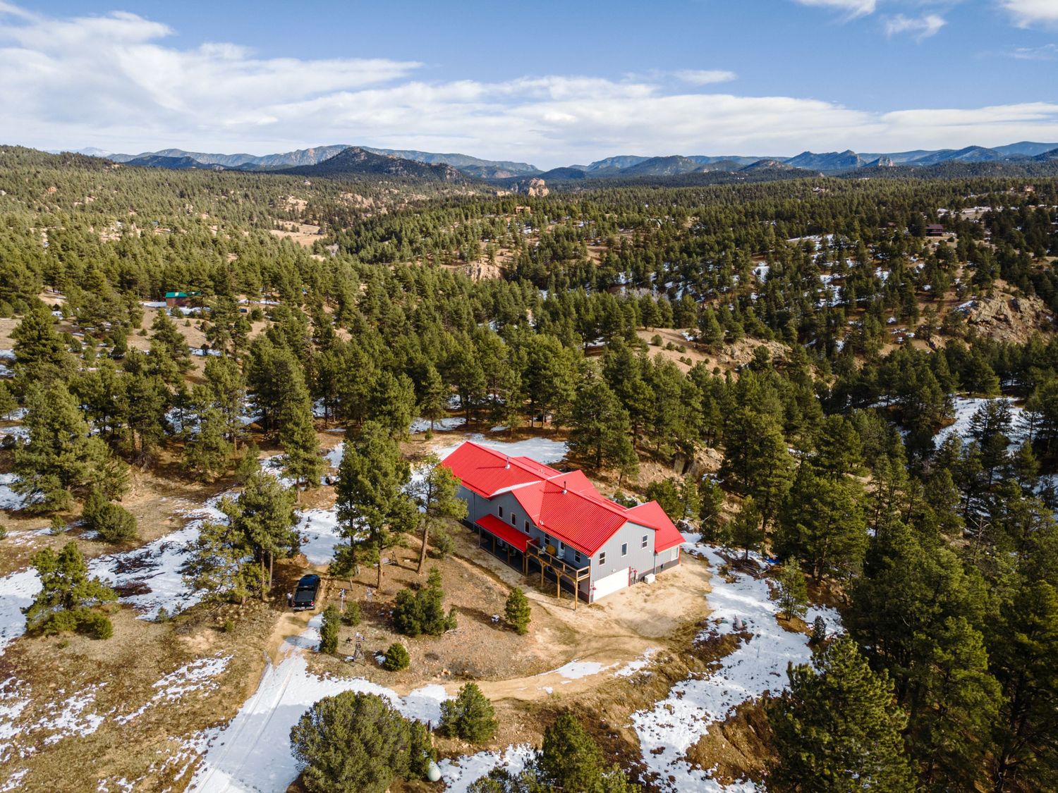 Aerial view of mountain property with red-roofed buildings nestled among pine trees and snow patches.