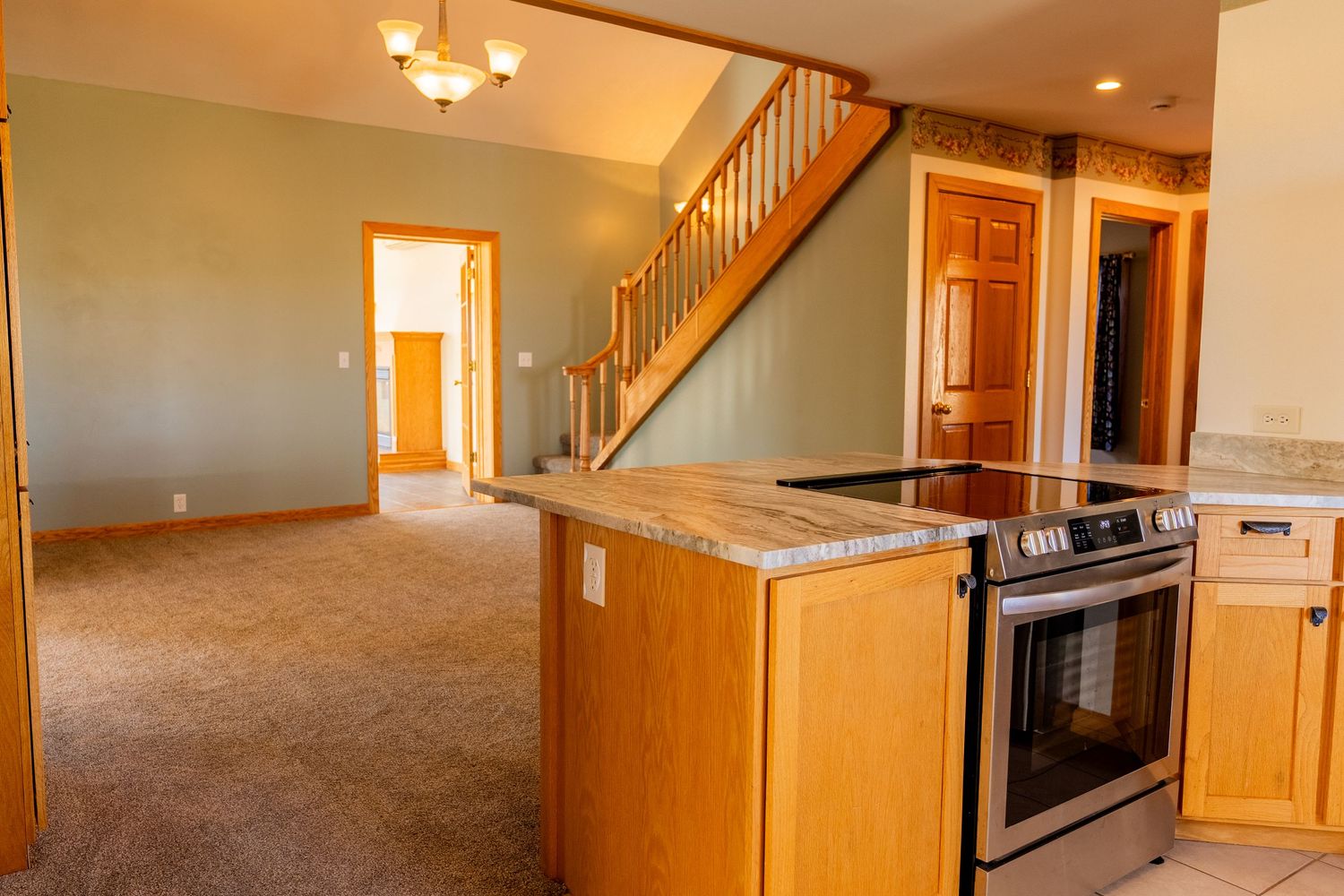 Open-concept kitchen with stainless steel range, granite counters, oak cabinets, and carpeted living area with staircase.