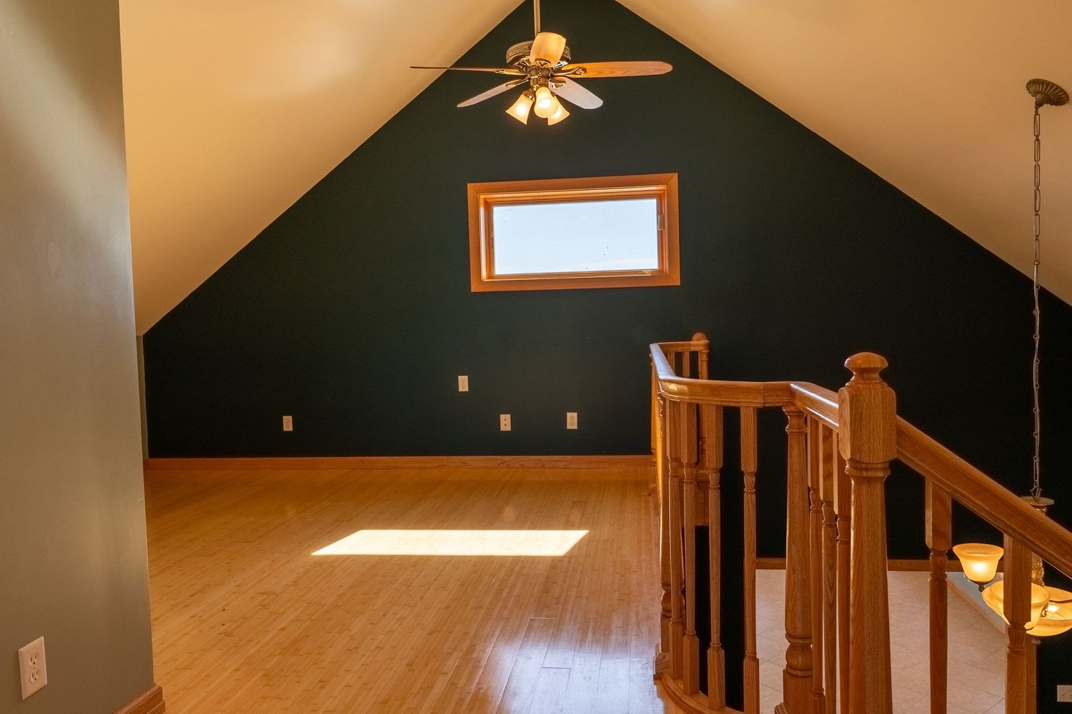 Loft space with vaulted ceiling, dark teal accent wall, hardwood floors, ceiling fan, and wooden staircase railing.