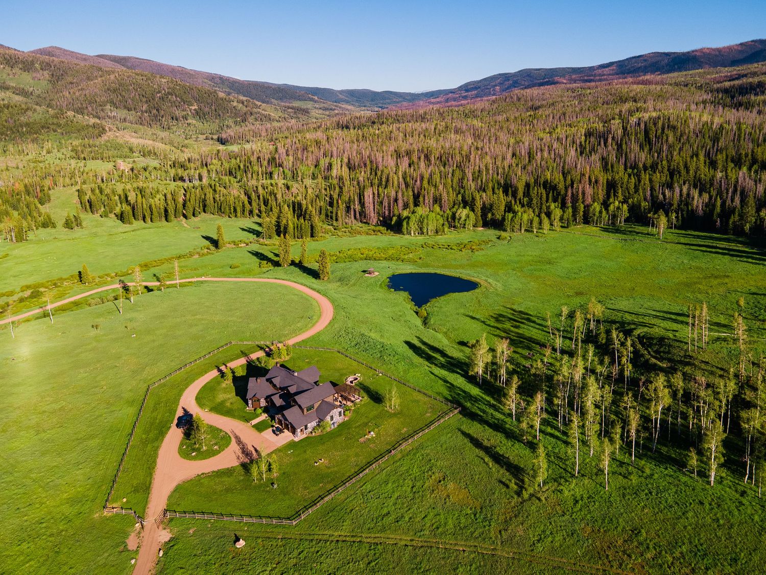 Aerial view of an exceptional Colorado mountain ranch with lush green meadows, a pond, and dense pine forests in a scenic valley.