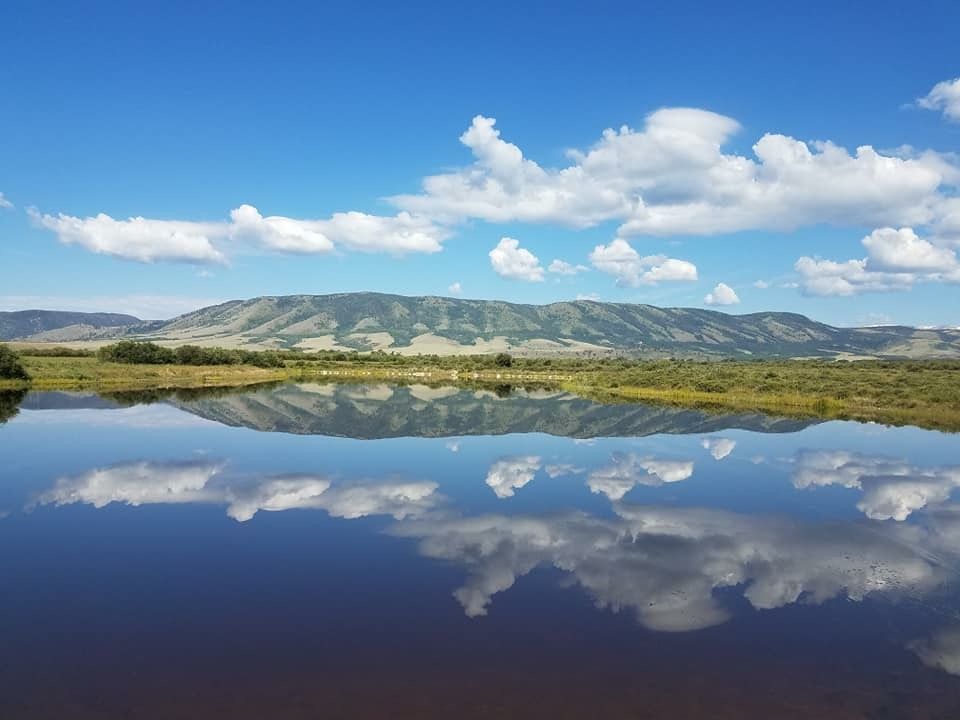 Serene Wyoming mountain lake reflecting blue sky and white clouds, with green hills in the background on a sunny day.