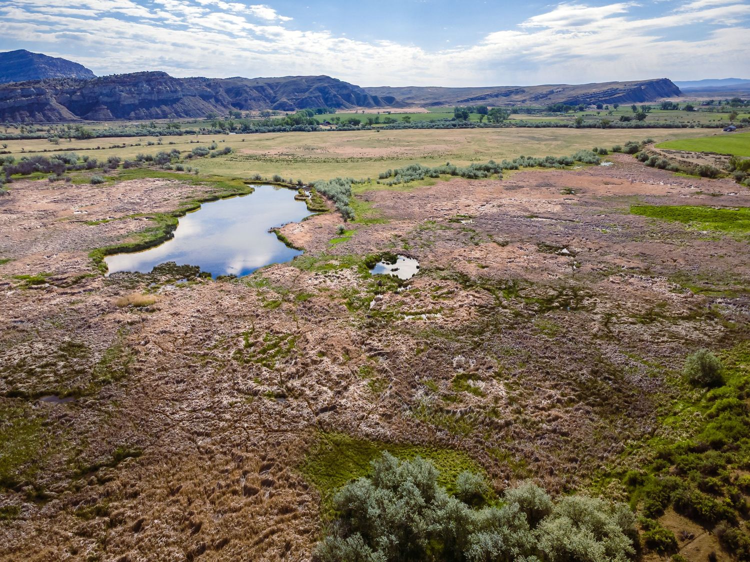 Aerial view of a small pond in an arid valley with rugged mountains, sparse vegetation, and dry terrain under cloudy skies.