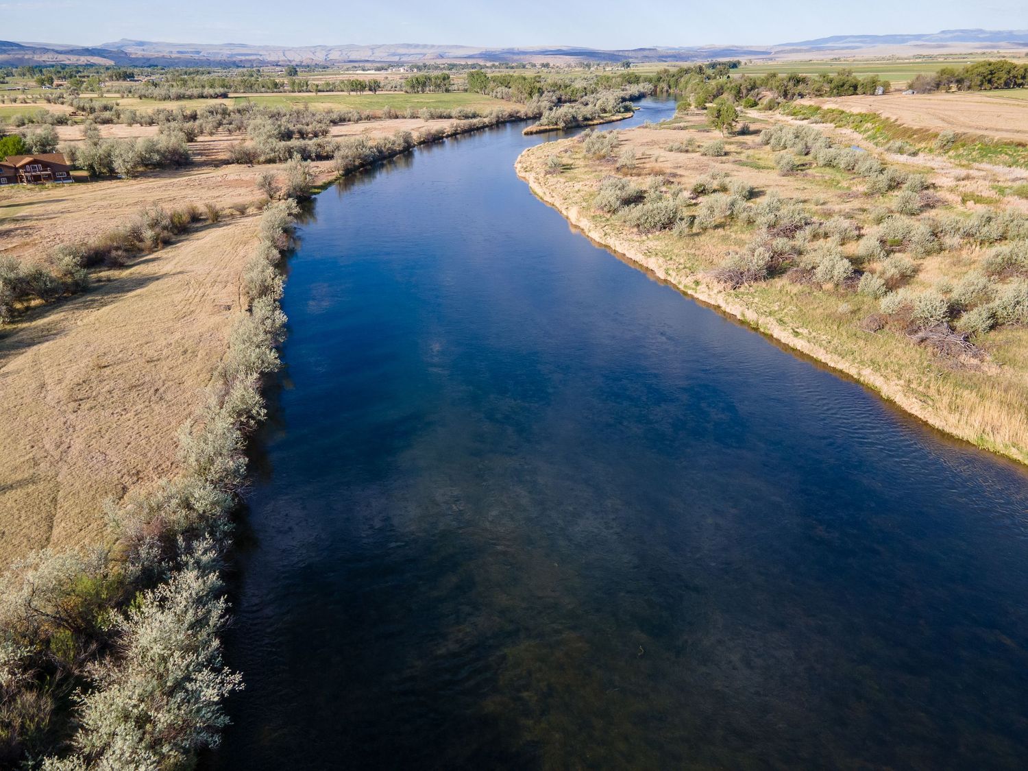 Aerial view of a calm wide river flanked by golden grasses and shrubs in open terrain.