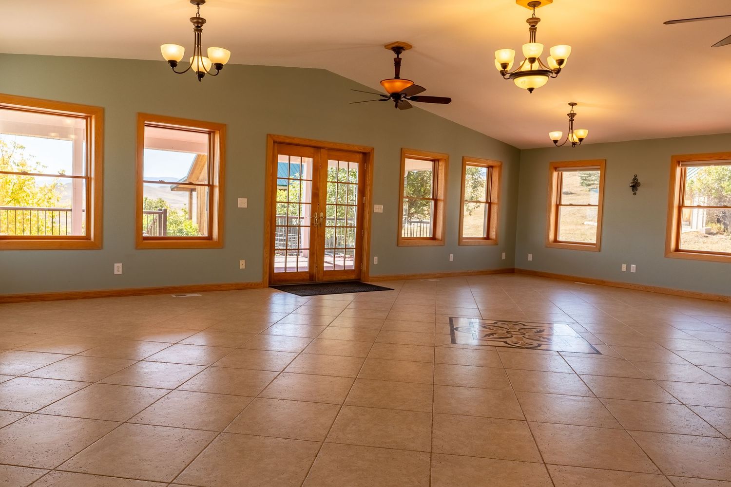 Spacious empty room with tile floors, ceiling fan, chandeliers, French doors, and large windows with wood trim.