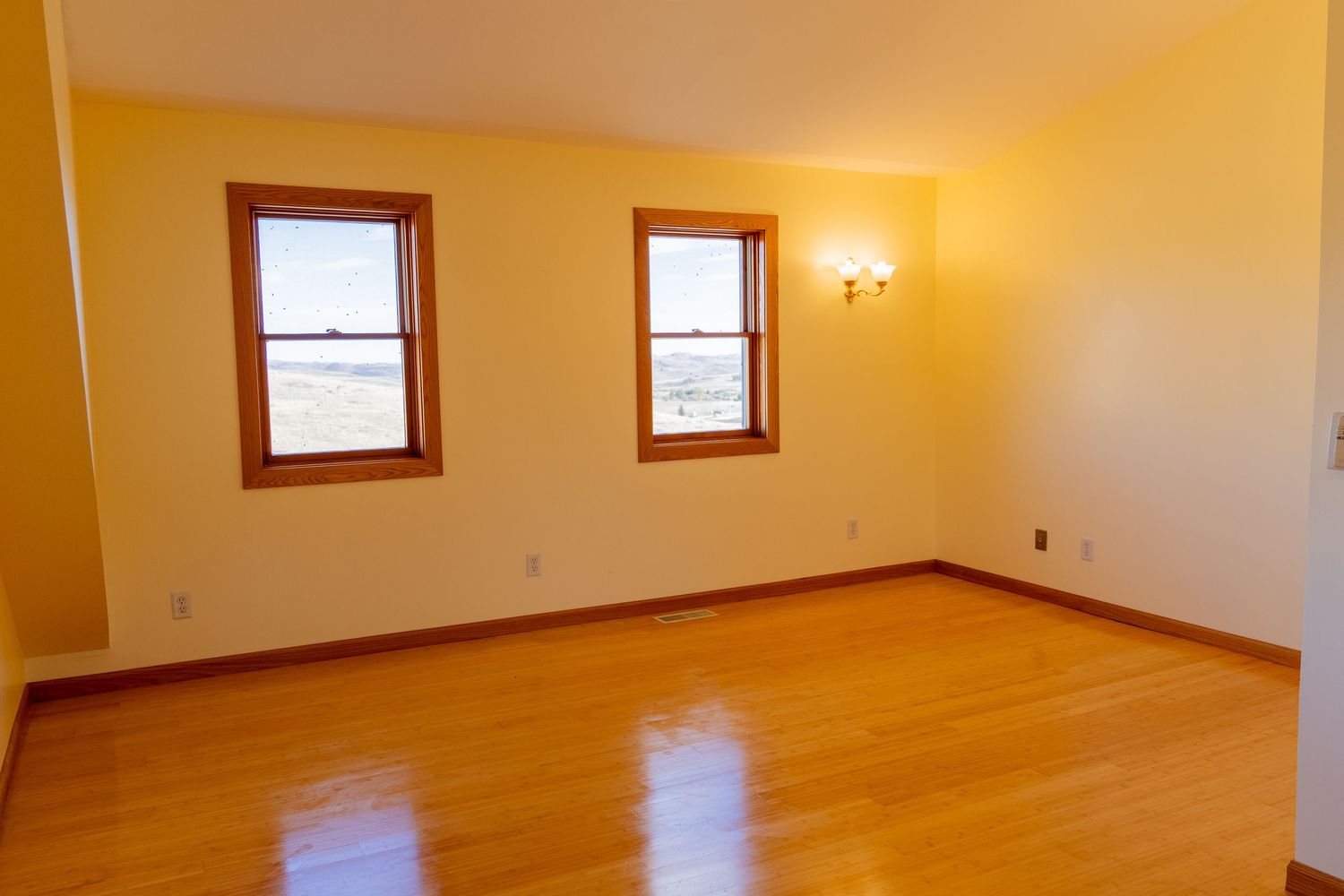 Empty room with hardwood floors, two windows, and warm yellow walls with wood trim.