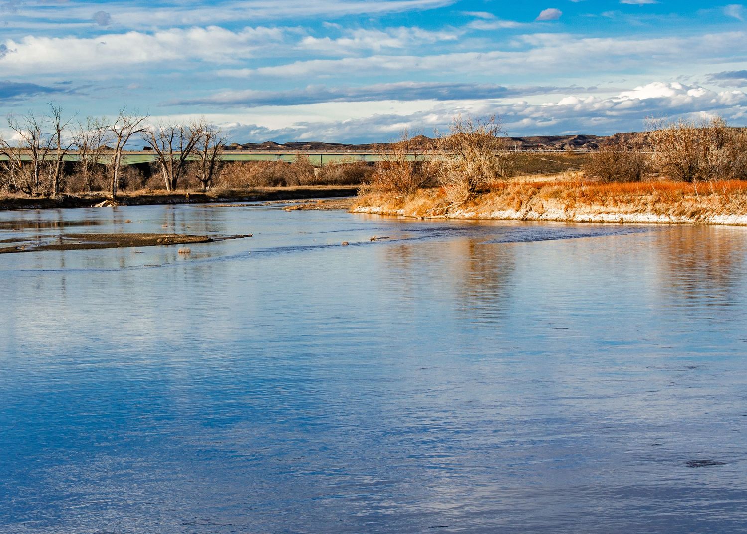 A serene winter river landscape with bare trees, golden banks, and blue sky reflections on calm water.