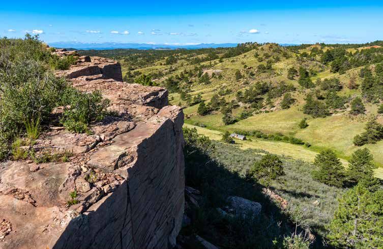 Rocky sandstone cliff edge overlooking green rolling hills and distant mountains under a bright blue sky.