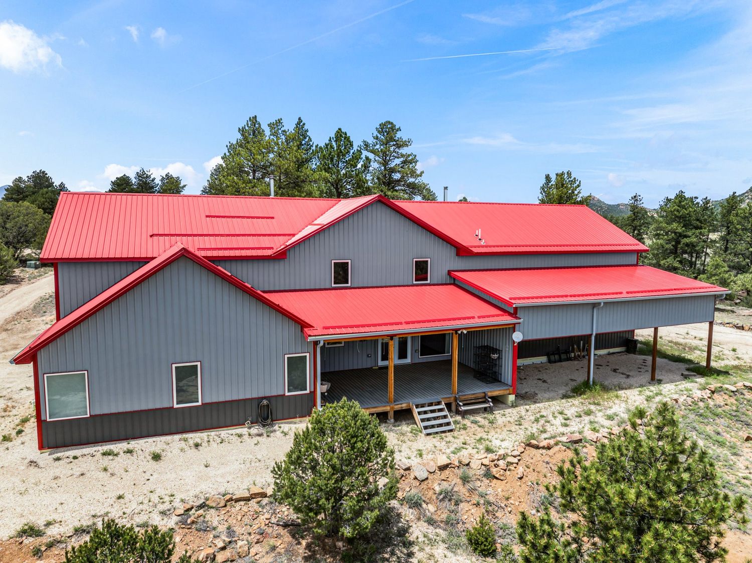 Large gray metal building home with bright red roof, covered porch, and pine trees in a rural setting.