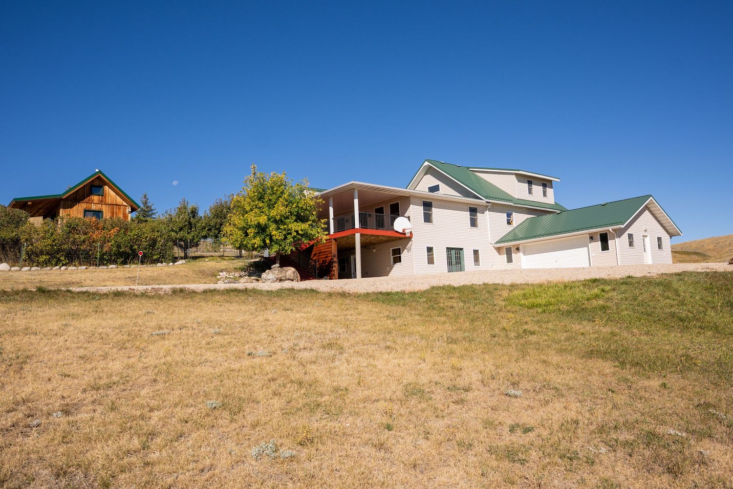 Rural property with large white farmhouse, red cabin, and green metal roof under clear blue sky on dry grassland.