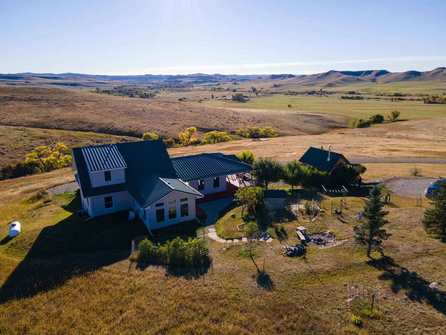 Aerial view of a rural farmhouse with blue roof surrounded by rolling hills and golden grasslands under a clear sky.