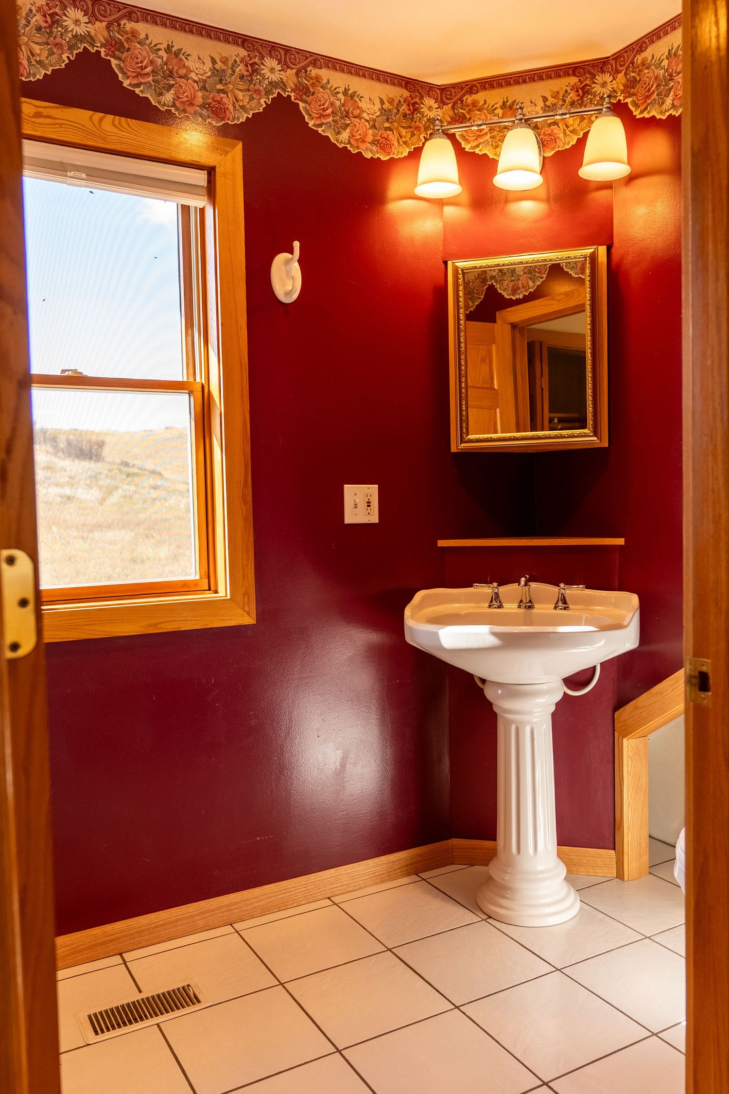 Elegant bathroom with deep red walls, white pedestal sink, gold-framed mirror, floral border, and tile flooring near a sunny window.