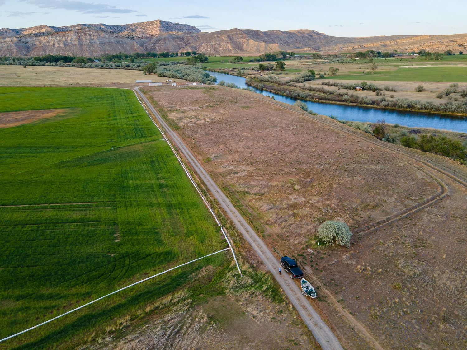 Aerial view of a winding river beside green farmland fields and a dirt road with mountains in the background.