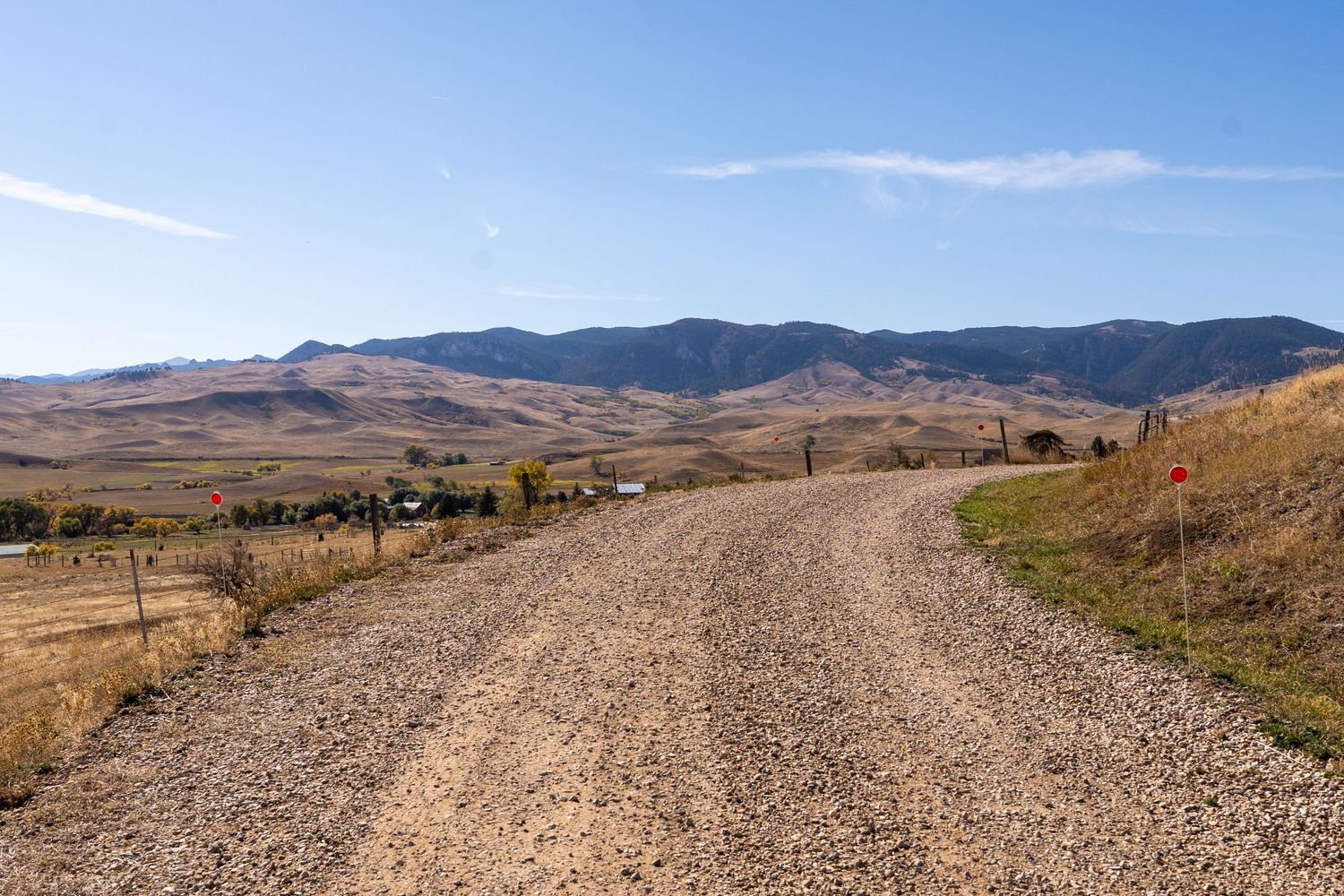 Gravel dirt road leading through open rural landscape with golden hills and mountain range under clear blue sky.