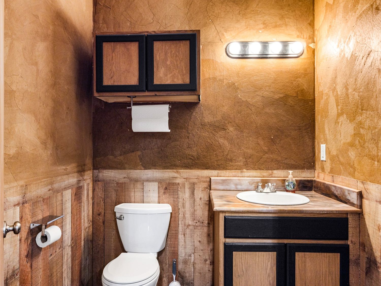 Rustic bathroom with wood wainscoting, faux-painted walls, white toilet, dark wood vanity, and wall-mounted cabinet with lighting.