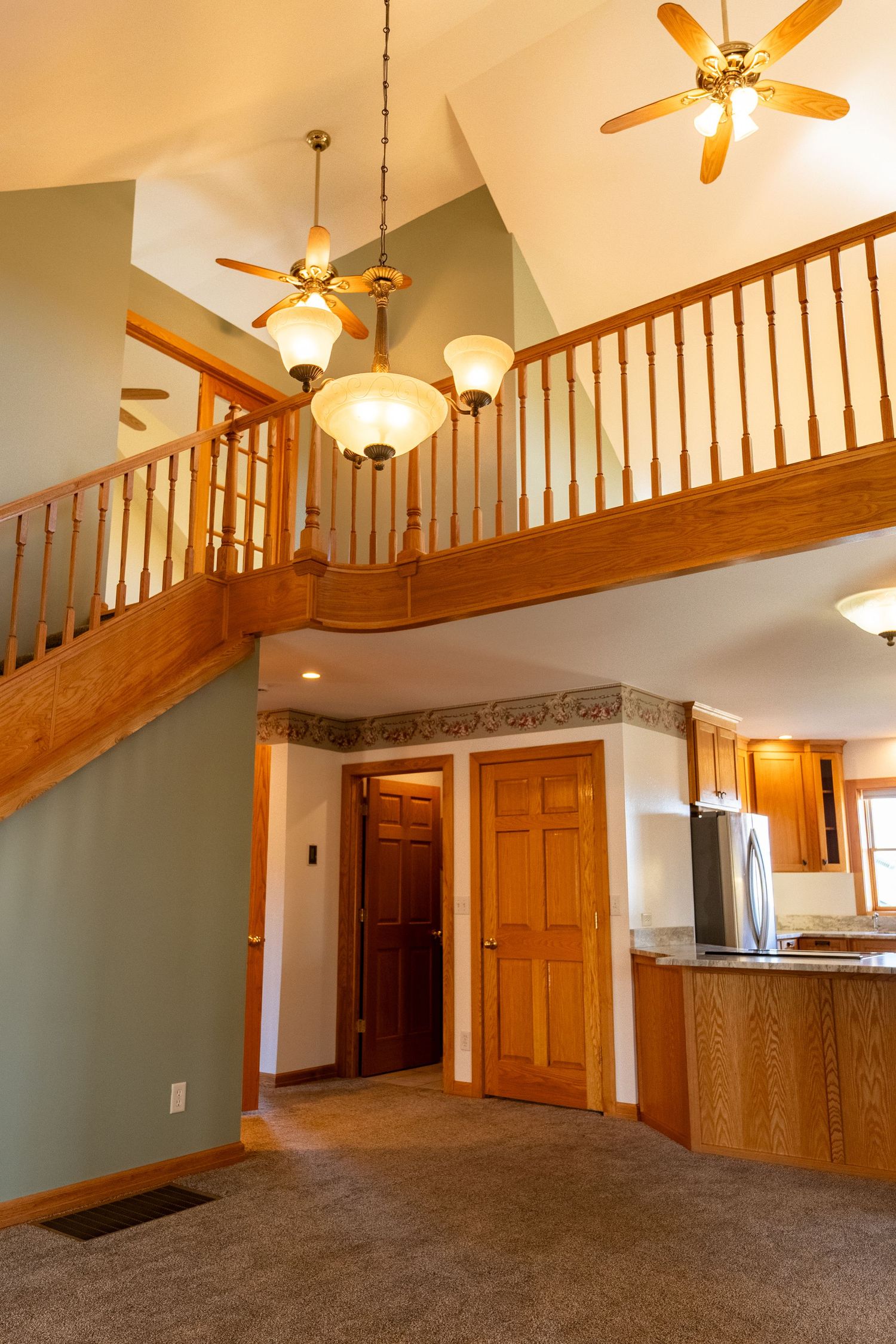 Open two-story home interior with oak staircase, ceiling fans, pendant lights, carpet floors, and kitchen with stainless refrigerator.