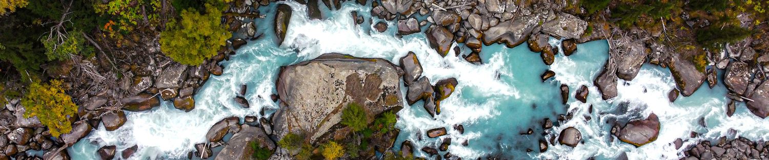 Aerial drone view of turquoise river rapids flowing through rocky terrain surrounded by autumn forest.