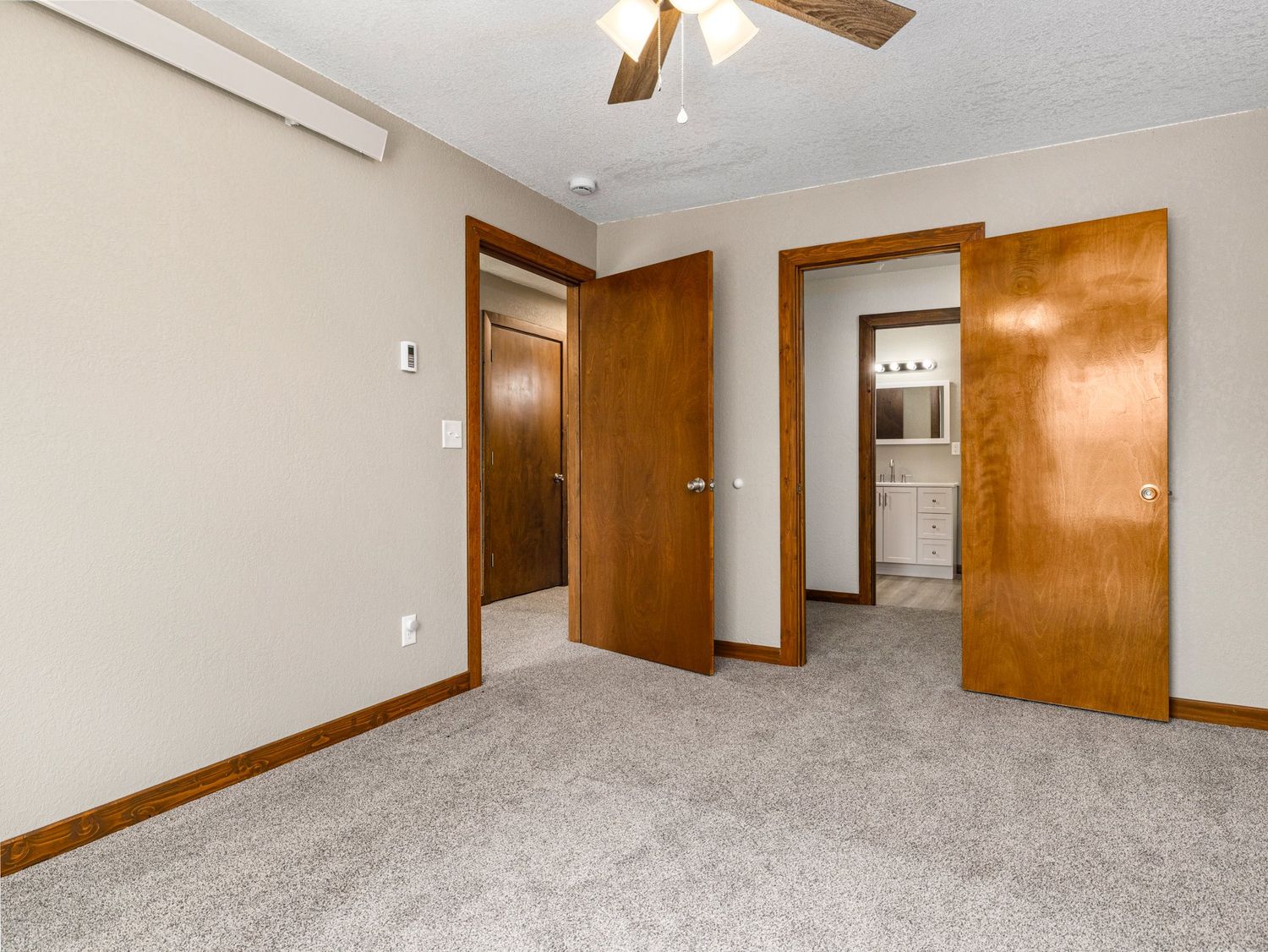 Empty carpeted bedroom with ceiling fan, two wooden doors leading to closet and ensuite bathroom, and neutral walls.