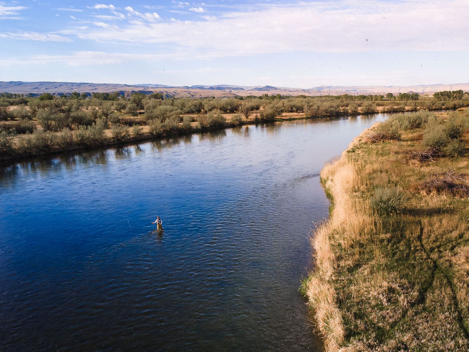 Aerial view of a lone paddleboarder on a calm blue river winding through golden grasslands under a partly cloudy sky.