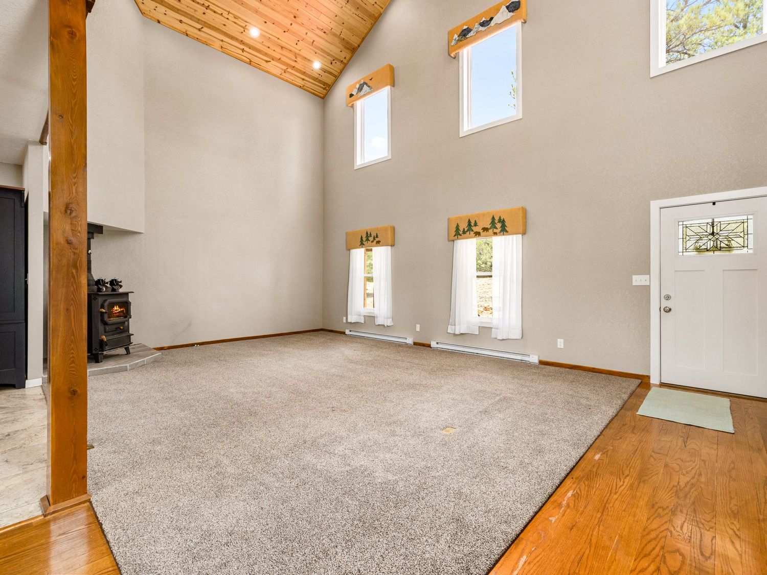 Spacious living room with vaulted wood ceiling, large windows, gray carpet, hardwood floors, and a wood-burning stove.