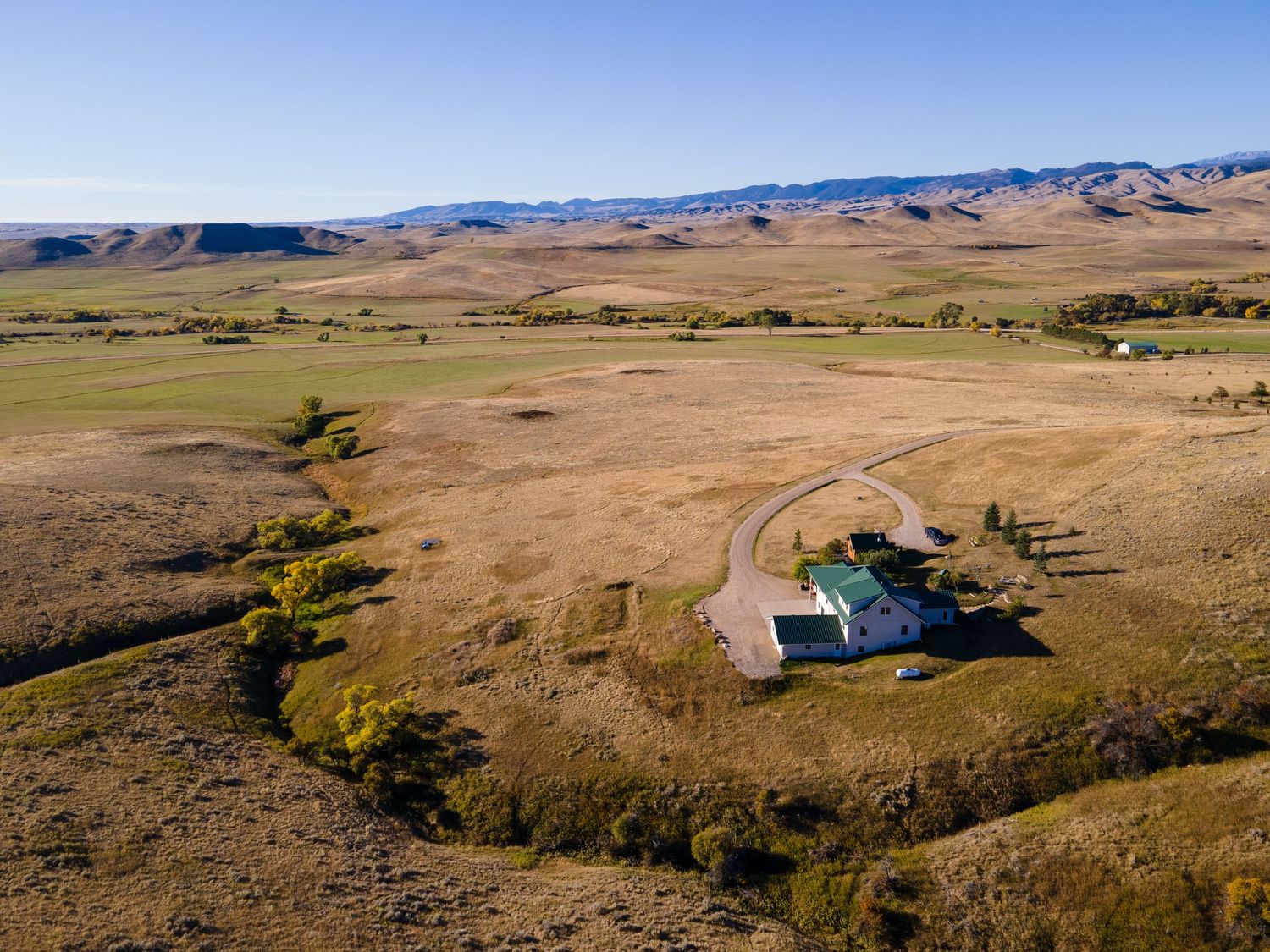 Aerial view of an isolated rural farmhouse surrounded by vast golden grasslands with mountains in the background under clear blue skies.