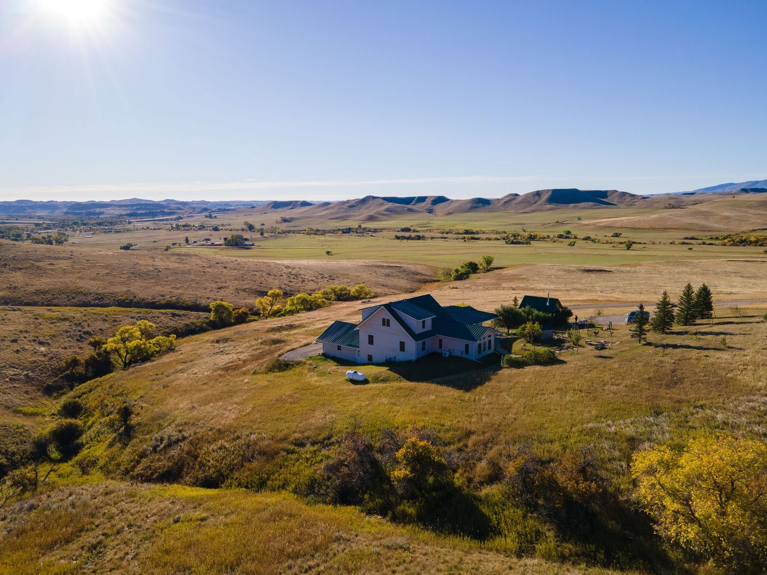 Aerial view of a rural farmhouse surrounded by rolling golden hills and open grasslands under a clear blue sky.