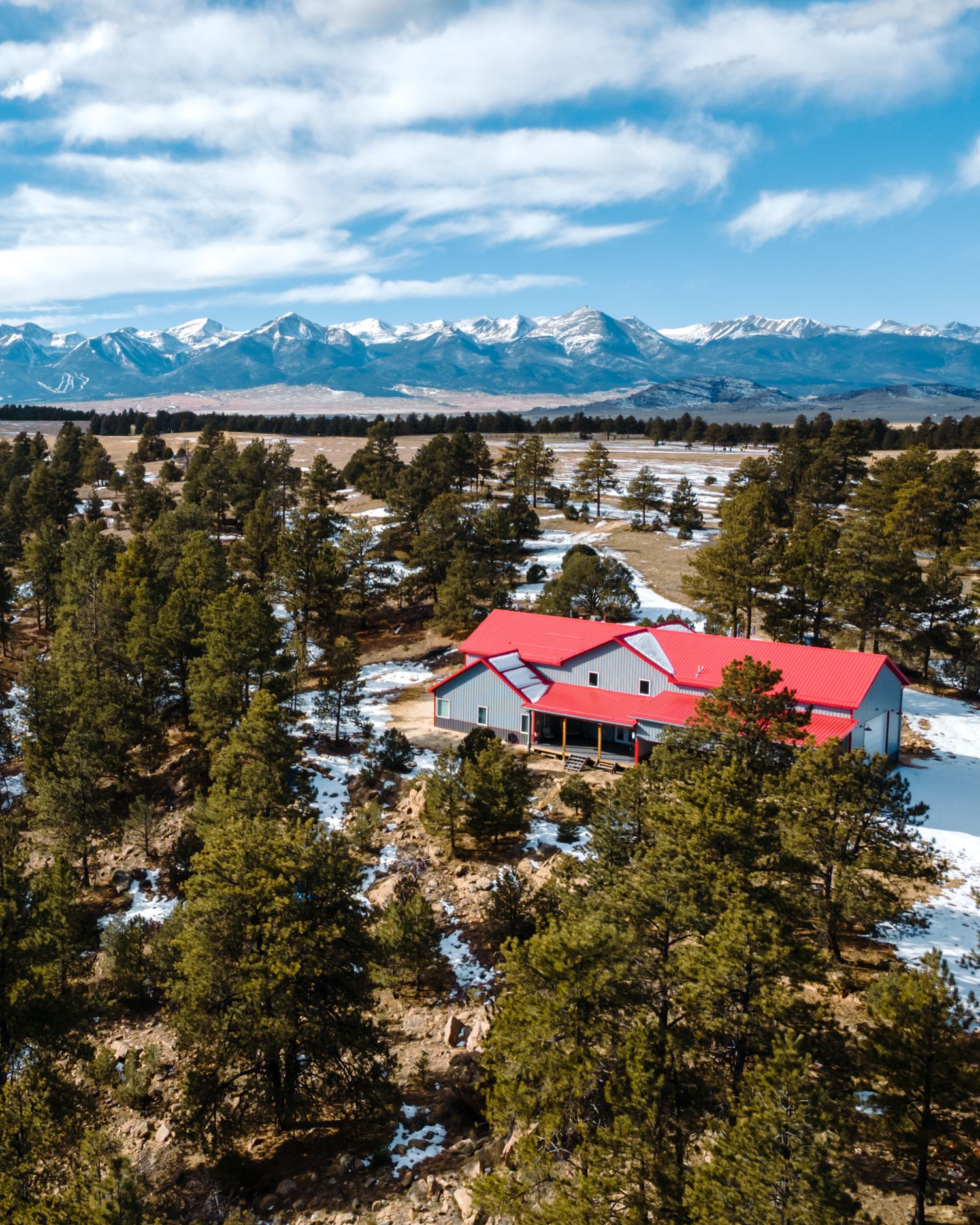 Snow-covered property with red-roofed buildings nestled among pine trees with mountain views.