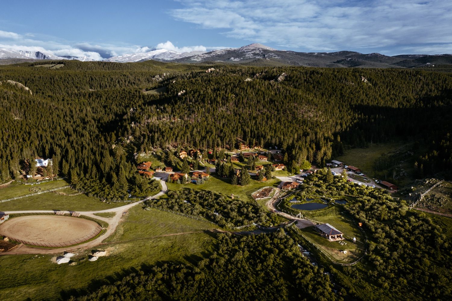Aerial view of a mountain resort nestled among dense evergreen forests with snow-capped peaks in the background.