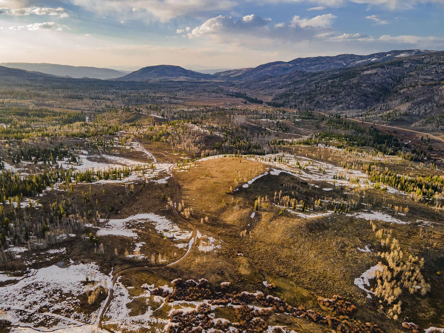Aerial view of a vast Colorado mountain valley with autumn trees, patches of snow, and winding paths under a partly cloudy sky.