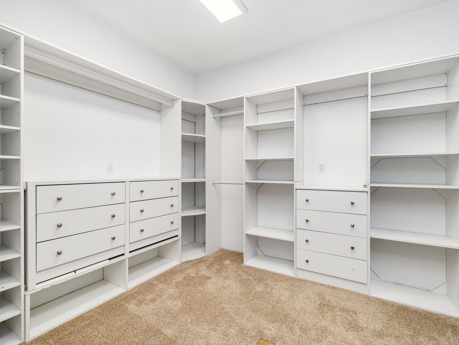 Spacious walk-in closet with white built-in shelving, drawers, and beige carpet flooring.