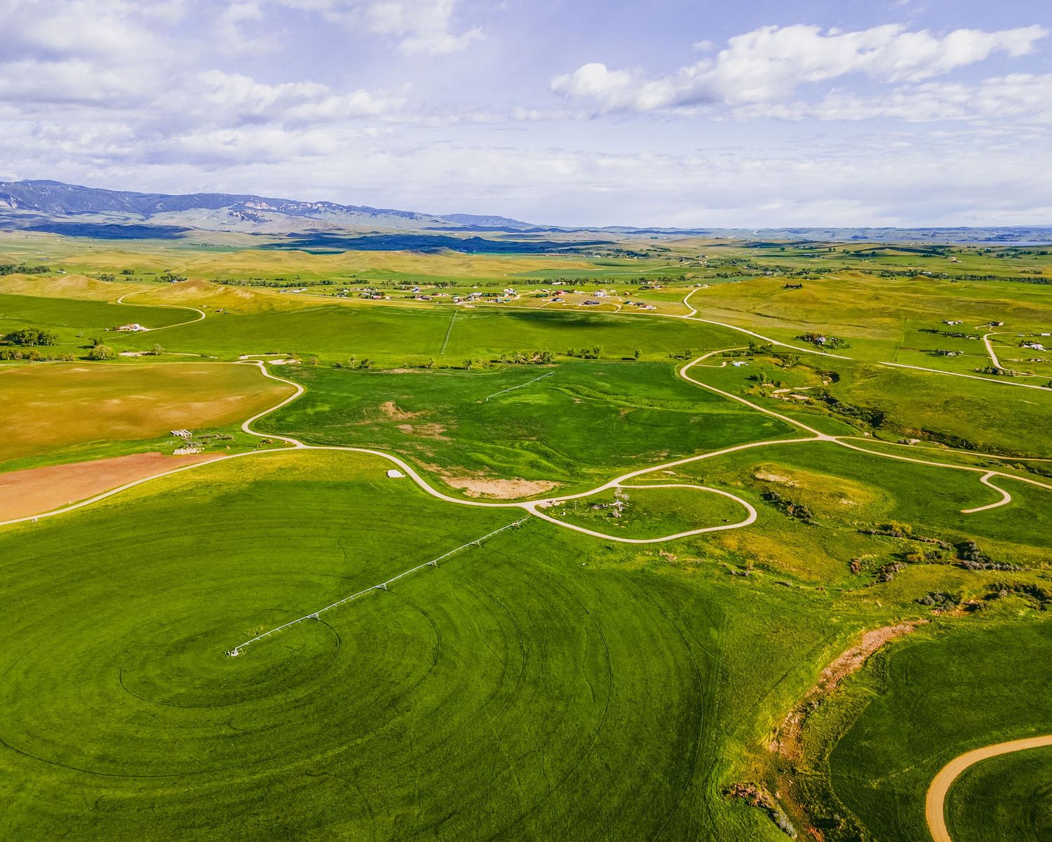 Drone shot of agricultural land showing curved pathways through green fields with distant mountain ranges.
