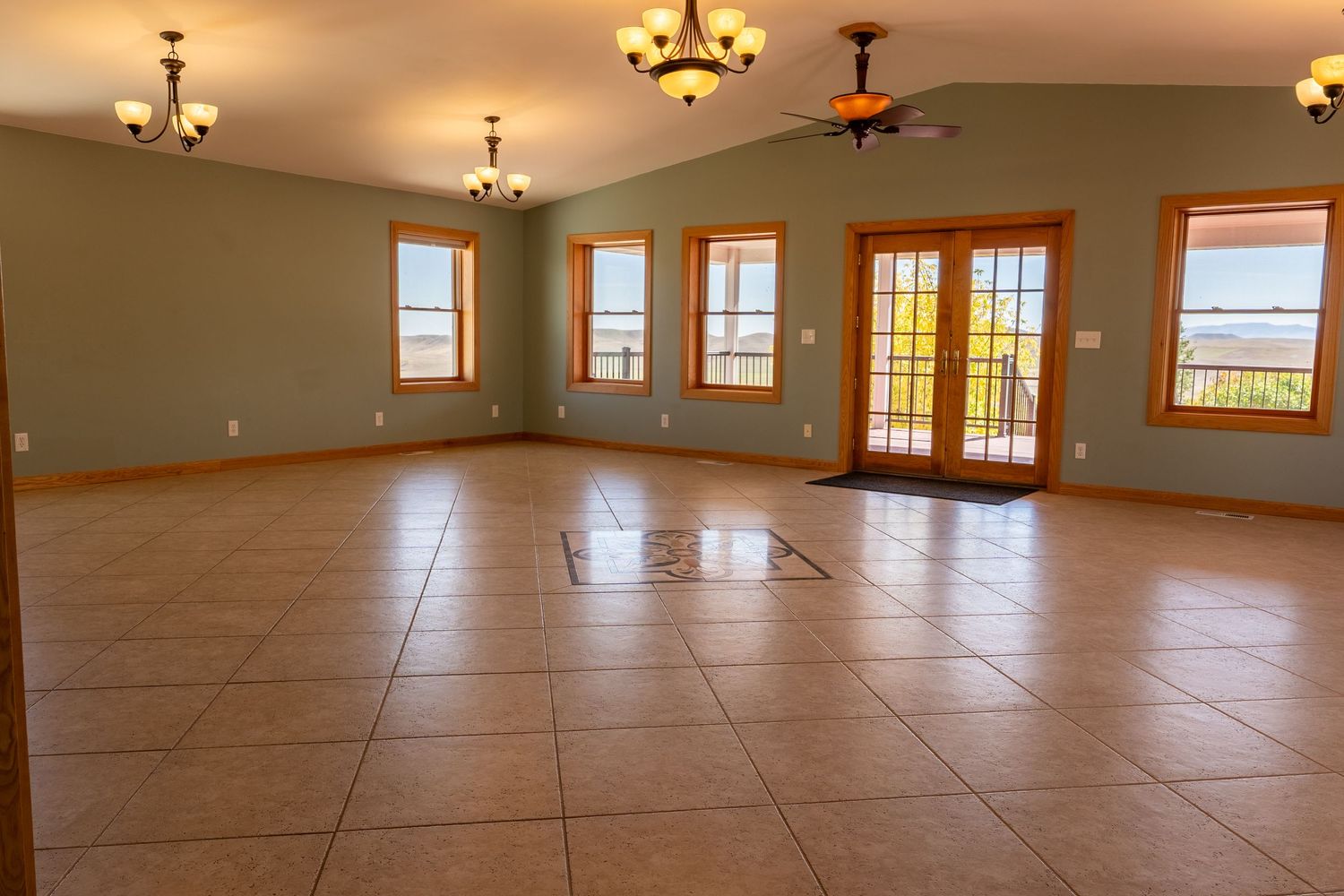 Spacious empty room with large tile floors, green walls, wood-trimmed windows, French doors, and elegant chandelier lighting.