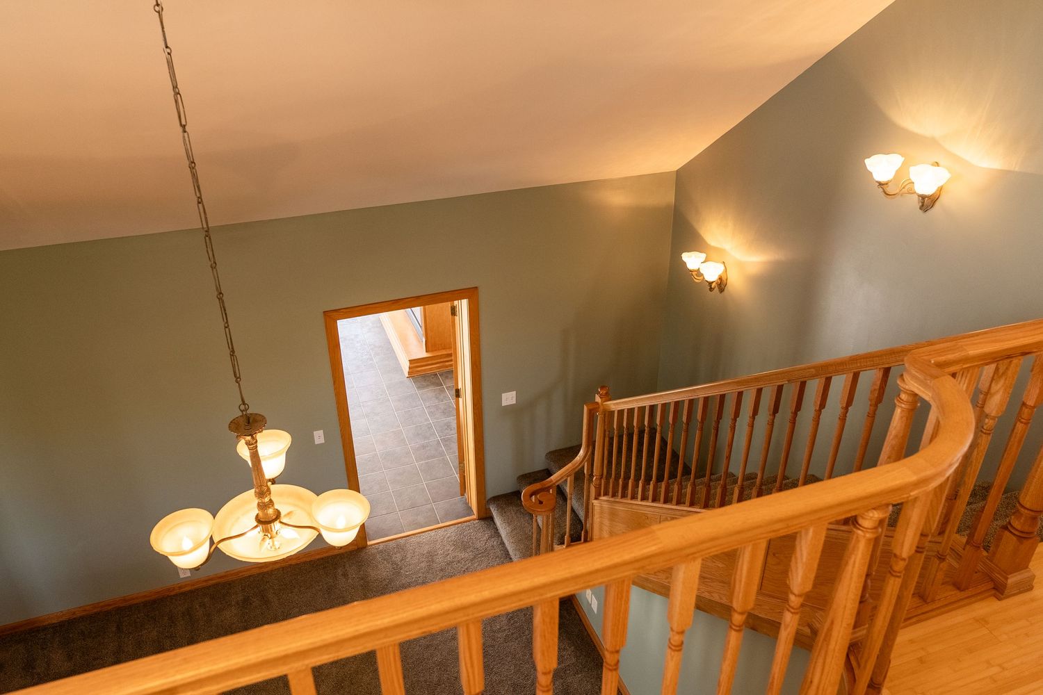 Curved wooden staircase railing with vaulted ceiling, chandelier, and wall sconces in a home upper hallway.
