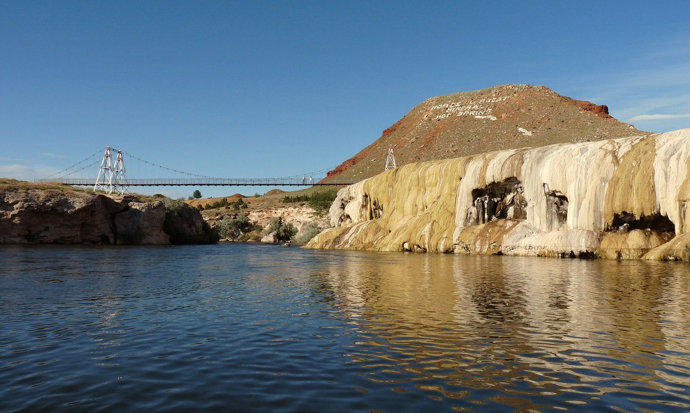Suspension bridge over calm river with white mineral deposits on red rock cliffs under clear blue sky.