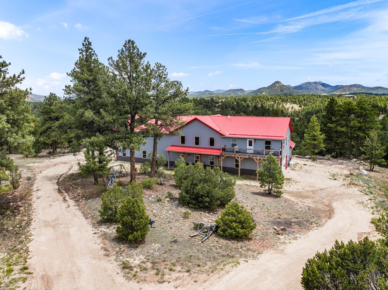 Aerial view of rural home with red metal roof surrounded by pine trees and mountain landscape.