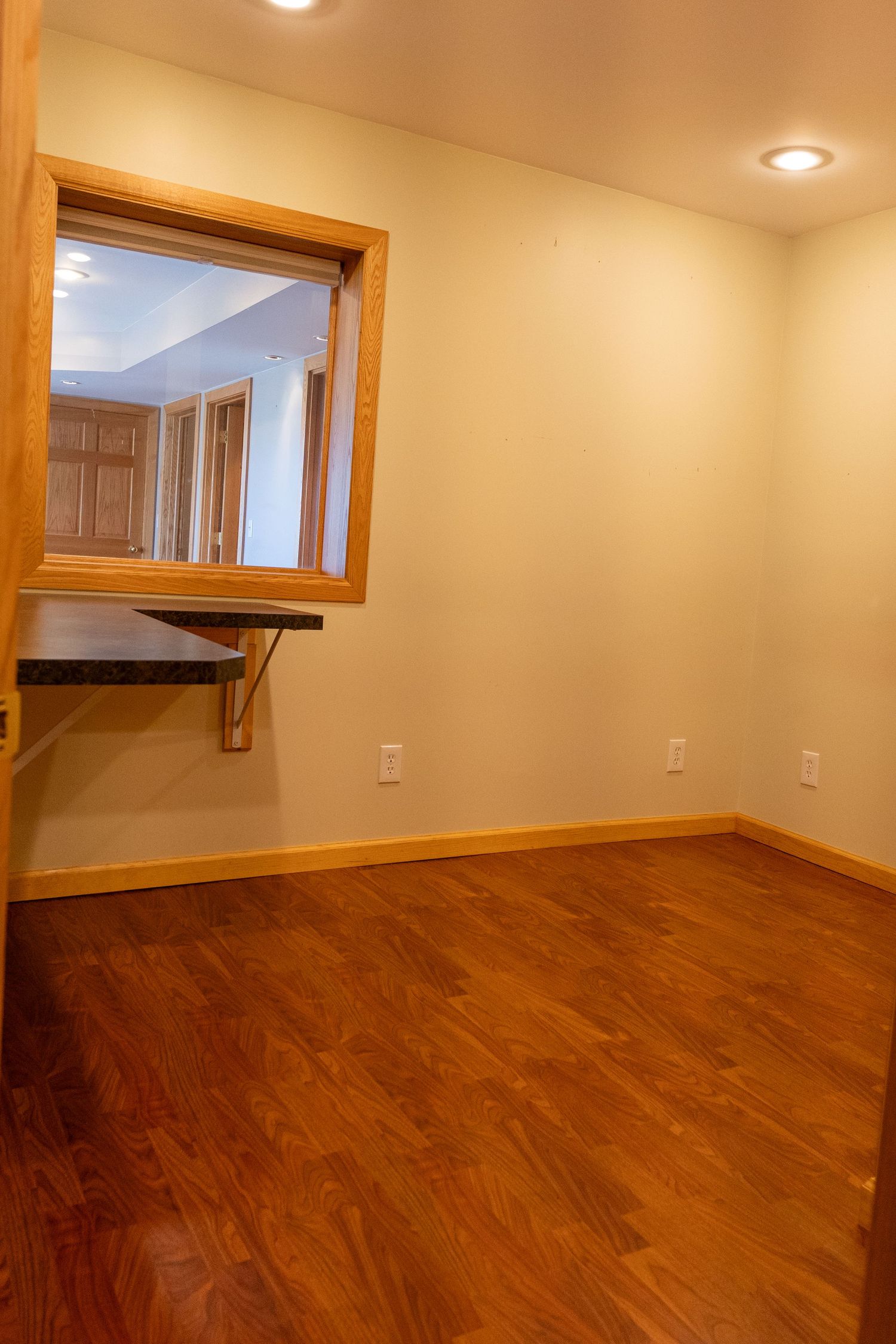 Empty room with hardwood floors, pass-through window, wall-mounted desk, recessed lighting, and neutral walls.
