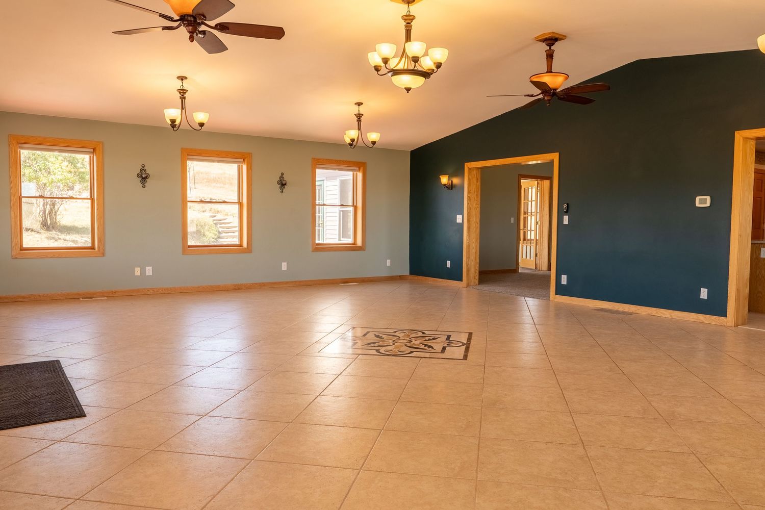 Spacious open-concept living room with tile floors, teal accent wall, vaulted ceilings, ceiling fans, and large windows.