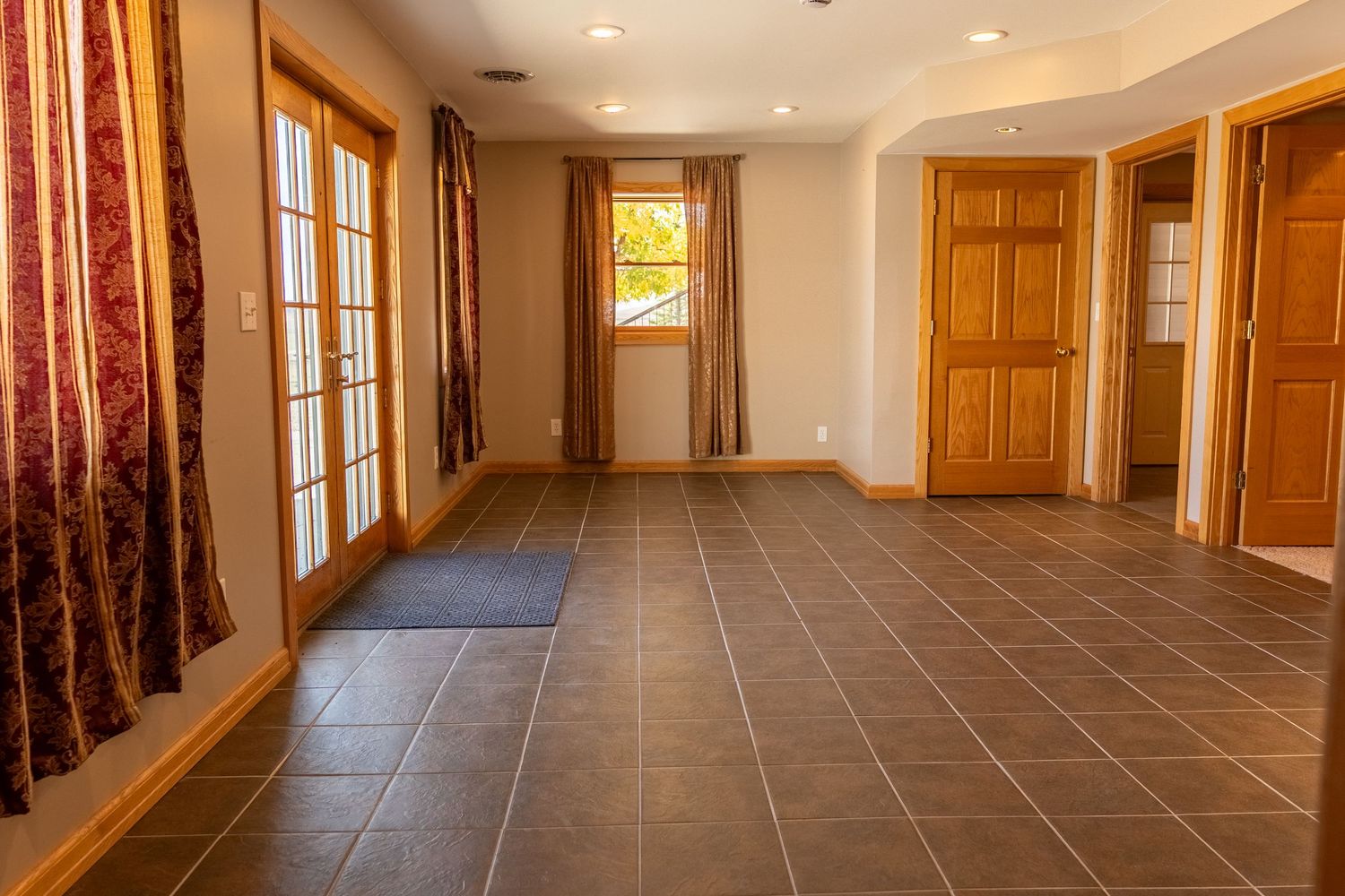 Spacious empty room with dark tile flooring, French doors, wood-trimmed windows, and multiple interior doors in neutral tones.
