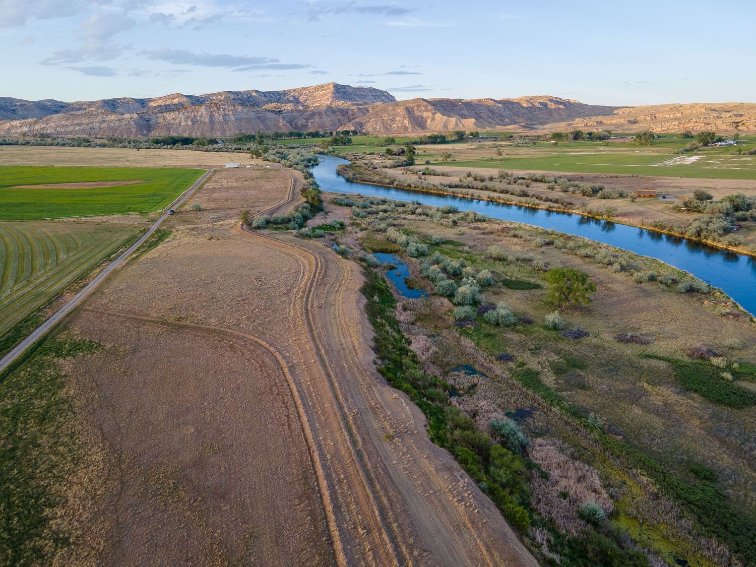Aerial panoramic view of a blue river winding through green farmland fields with mountain ridges at golden hour.