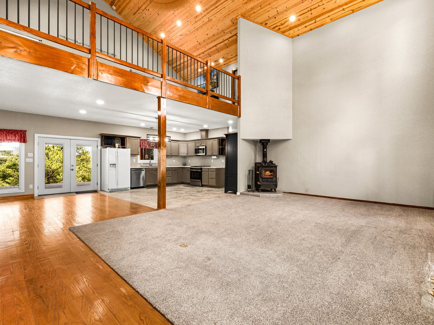 Spacious open-concept living room with hardwood floors, carpet area, wood stove, high ceilings, loft, and modern kitchen in background.