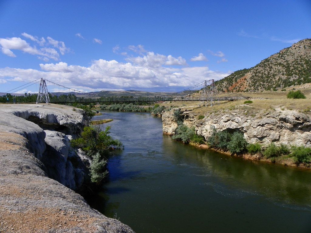 A scenic river flowing through rocky terrain with green hills, sparse vegetation, and a suspension bridge under a partly cloudy blue sky.