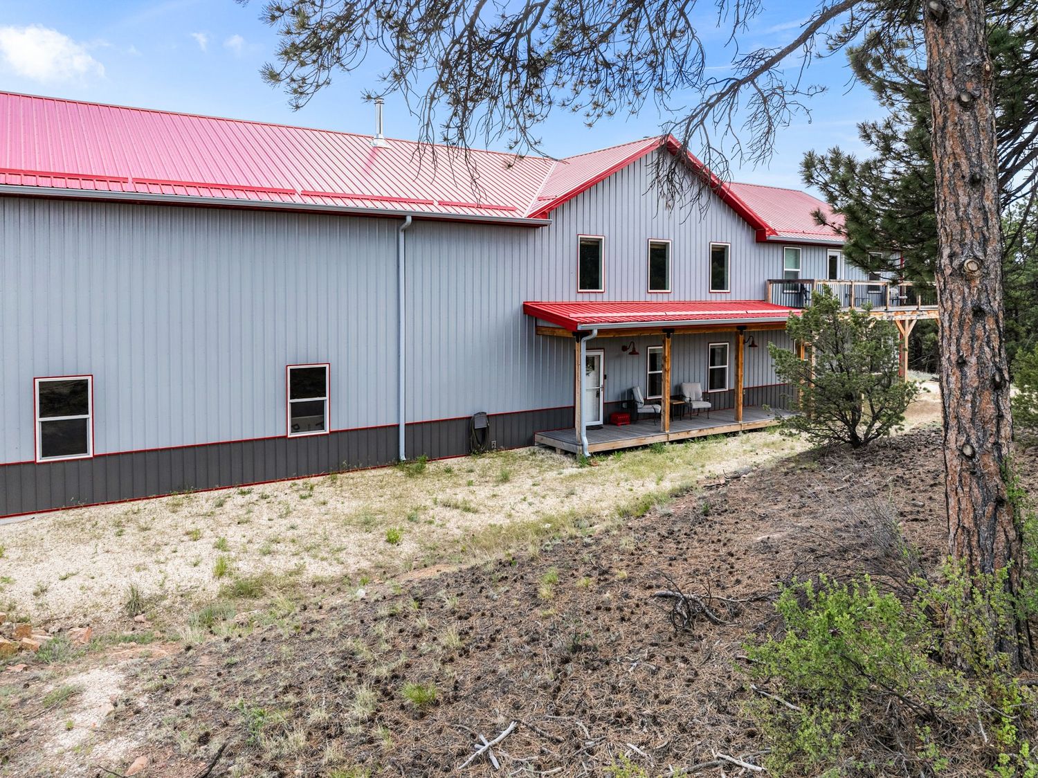 Gray metal barn-style building with red roof and covered porch entrance surrounded by pine trees and sandy landscape.