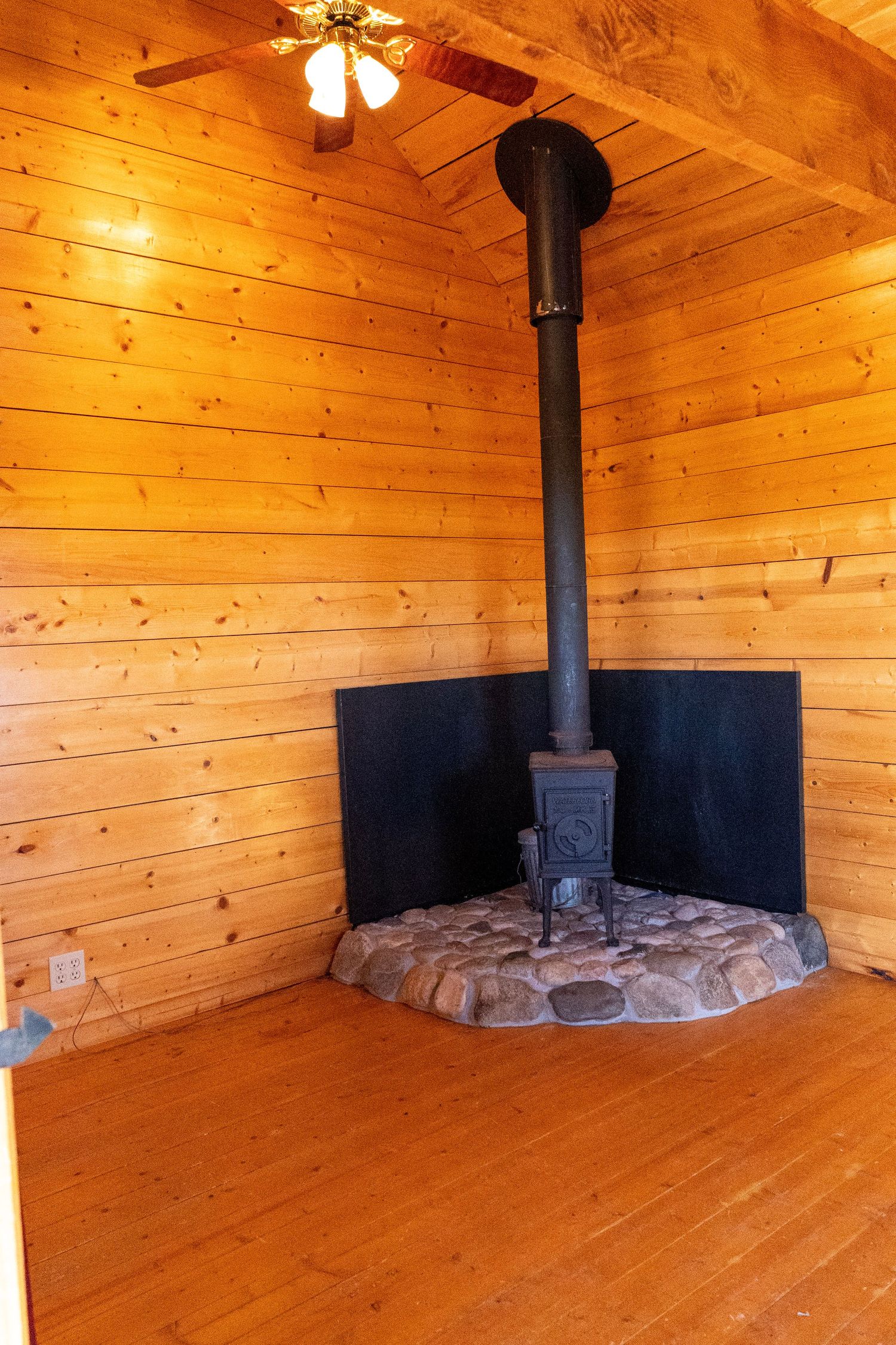 Cozy wood-paneled cabin interior with a cast iron wood-burning stove on a stone hearth and black chimney pipe.